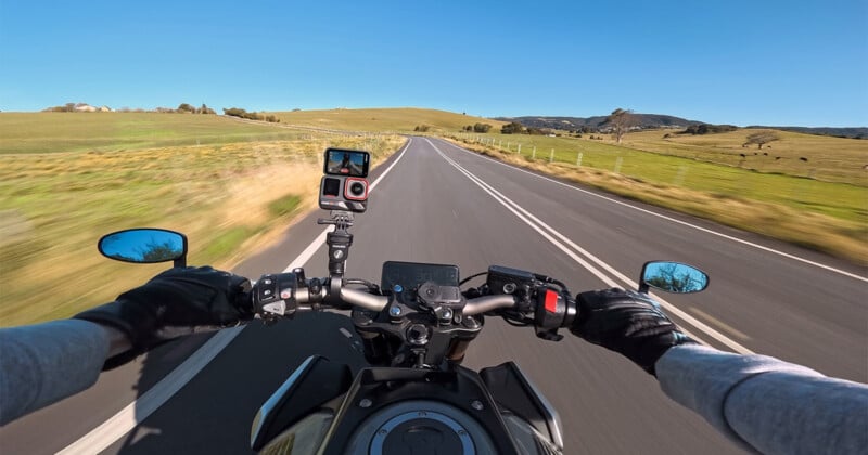 Point-of-view photo of a motorcyclist riding on an open road. The road is flanked by green fields under a clear blue sky. The rider's gloved hands are visible on the handlebars, with a camera mounted on the bike capturing the journey.