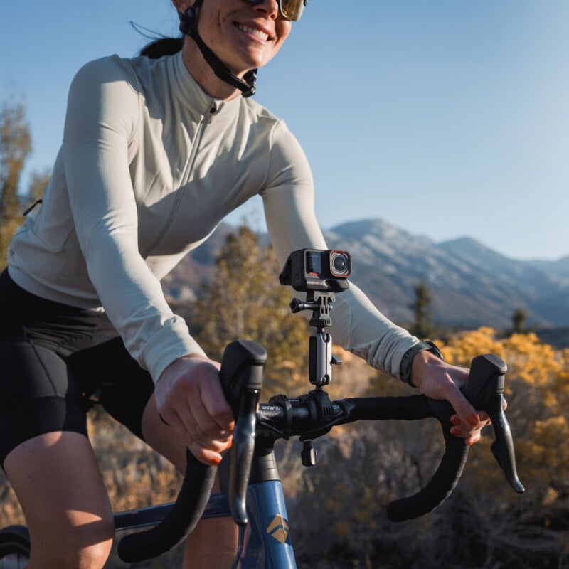 A person wearing a white jacket and sunglasses rides a bicycle with a camera mounted on the handlebars. In the background, there are trees and snowy mountains under a clear blue sky.