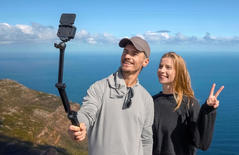 A man and woman smiling while taking a selfie on a cliffside. The man is holding a selfie stick, and the woman is making a peace sign. The background features a scenic ocean view under a clear blue sky.