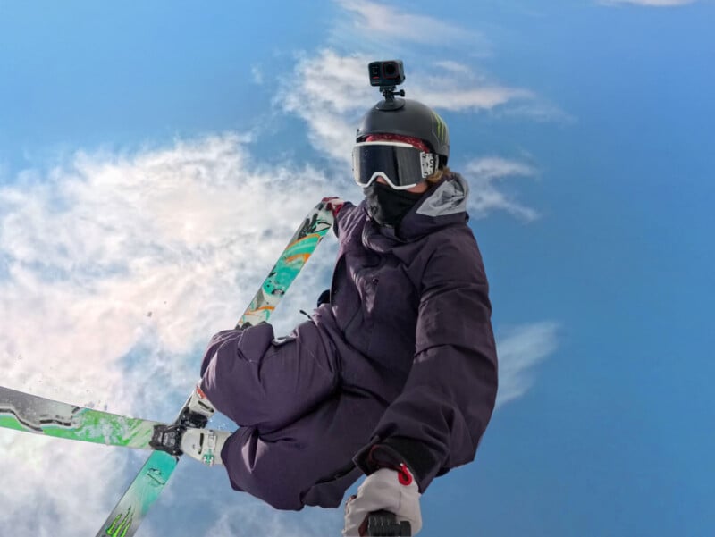 A skier in a purple outfit performs a mid-air trick against a bright blue sky. They wear a helmet with a mounted action camera and goggles, while one ski is visible. White clouds form a partial backdrop.