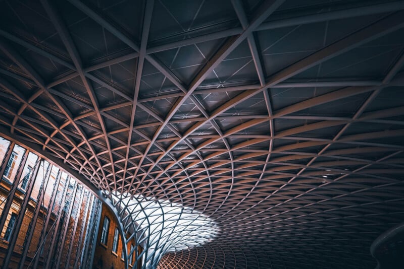 A modern architectural ceiling with a geometric, crisscrossed pattern made of metal beams and glass. Soft light filters through, illuminating the space with a warm glow. A curved wall with vertical windows can be seen on the left side.