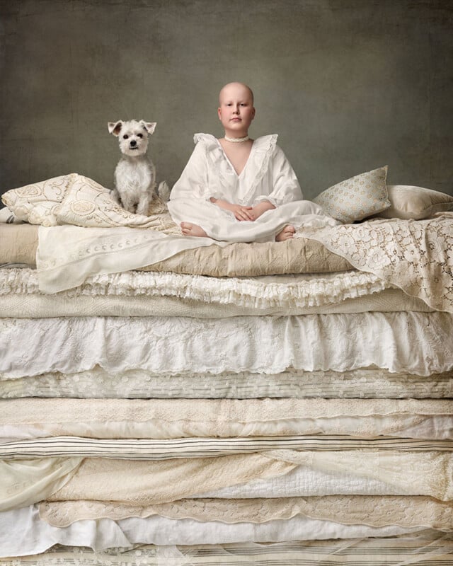 A person in a white gown sits atop a tall stack of mattresses, alongside a small dog. The scene resembles a fairy tale setting, with intricately patterned linens creating a whimsical atmosphere.