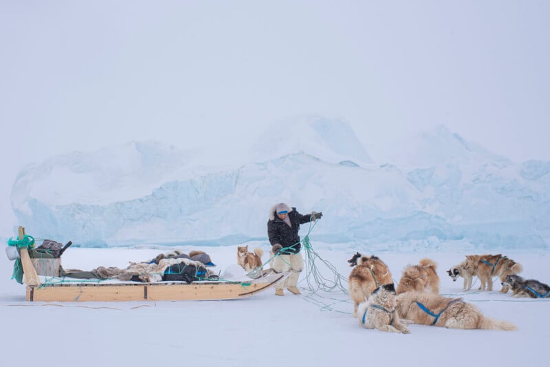 A person stands on a snowy landscape, tending to a sled with several dogs harnessed. An icy, mountainous backdrop completes the scene, with a cloudy sky above. The person is dressed in heavy winter clothing, suggesting a cold environment.