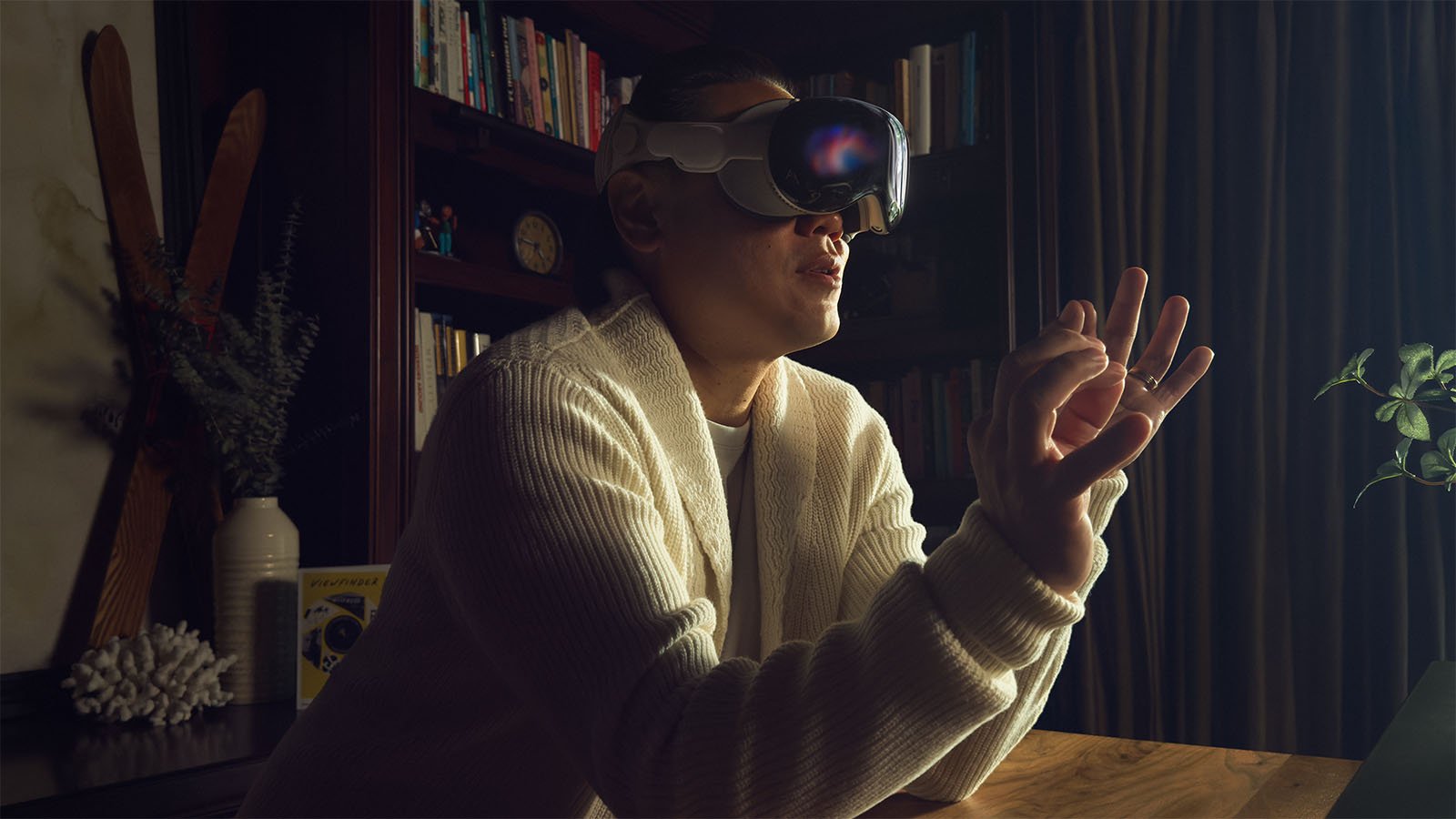 A person wearing a VR headset gesturing with their hand while seated at a wooden table. They are in a room with a bookshelf in the background, partially illuminated by light.