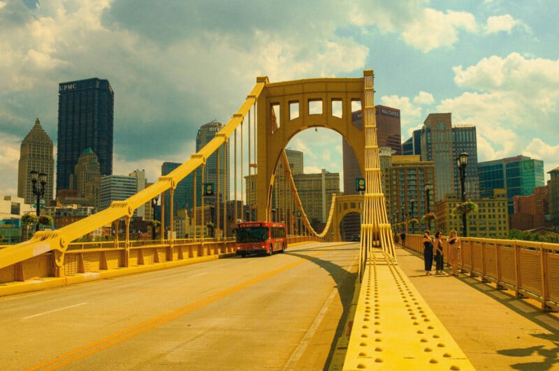 A yellow suspension bridge with a red bus crossing it, set against a city skyline of tall buildings under a cloudy sky. Two people stand on the bridge walkway, facing the cityscape.