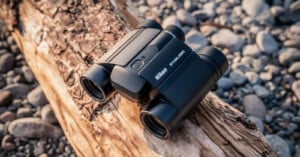 Close-up of a pair of Nikon binoculars resting on a rough piece of driftwood, with a background of pebbles. The binoculars are black and compact, displaying the brand name clearly. Natural lighting provides a warm tone to the scene.