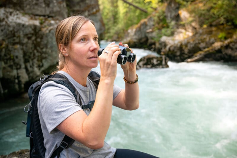 A person with a backpack is sitting by a flowing river, looking through binoculars. The setting is a rocky area with lush green forest in the background. The person is wearing a gray t-shirt and appears focused on observing something.