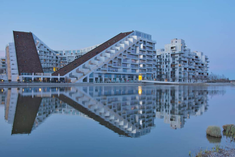 A modern architectural complex with a unique sloped roof is reflected in the calm water of a nearby lake. The sky is clear and blue, enhancing the serene atmosphere.