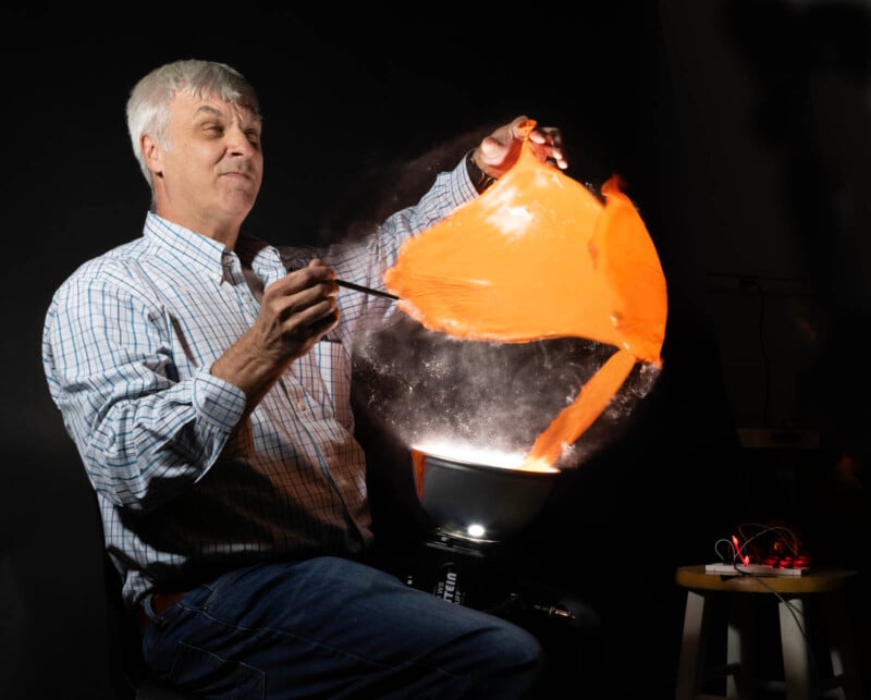 A man in a plaid shirt sits on a stool against a dark background. He is bursting an orange balloon with a black stick, revealing a splash effect. A small device is on another stool nearby.