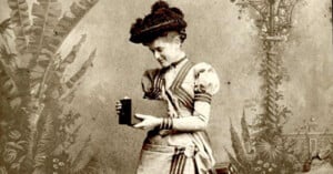 A vintage photograph of a woman in an elaborate dress and hat, holding a book and looking at it. The background is ornate with floral and architectural designs, typical of late 19th-century photography.