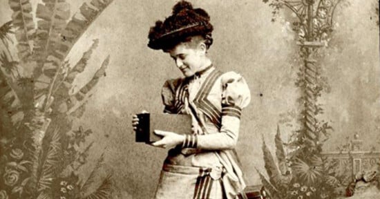 A vintage photograph of a woman in an elaborate dress and hat, holding a book and looking at it. The background is ornate with floral and architectural designs, typical of late 19th-century photography.