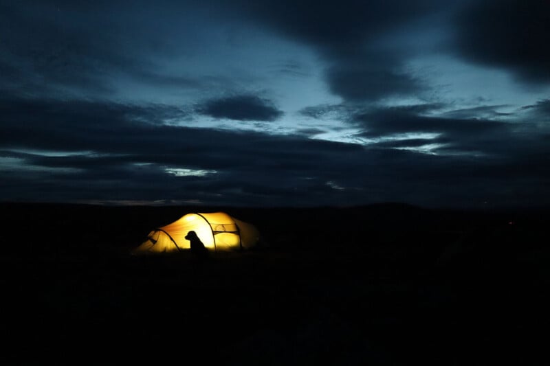 A glowing tent is illuminated from within against a dark, cloudy evening sky. The silhouette of a person is visible near the tent, creating a serene and solitary nighttime camping scene.