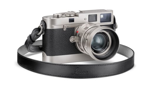 A Leica camera with a silver body and a black textured grip, featuring a large lens and a black leather strap embossed with "Leica." The camera is angled to show both the lens and the top dials with a vintage aesthetic.