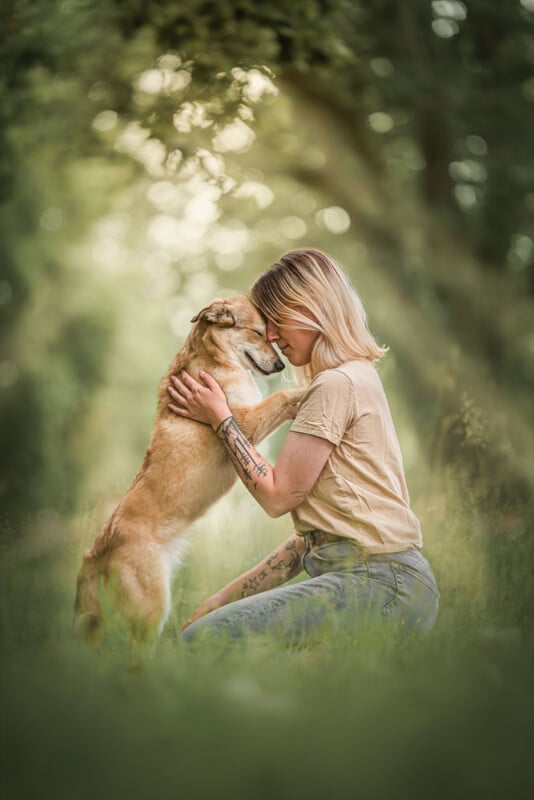 A person with short blonde hair kneels in a lush green setting, gently embracing a tan dog. They touch heads affectionately, surrounded by soft-focus greenery, creating a serene and intimate moment.