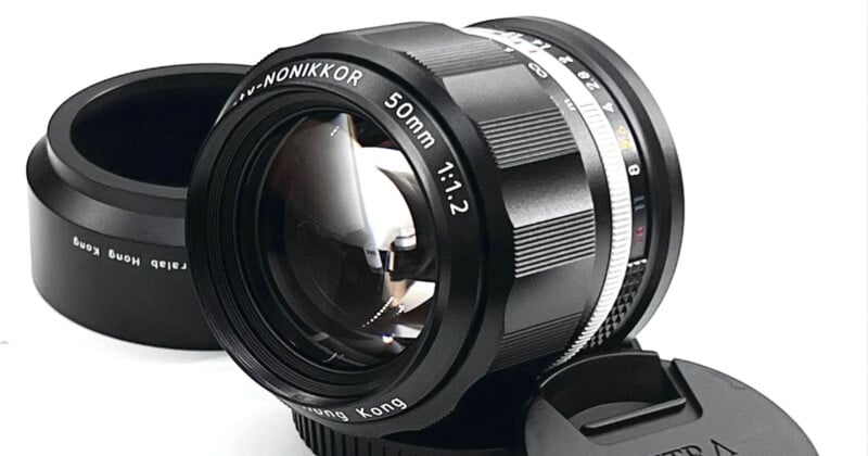 A close-up of a black Nikon 50mm 1:1.2 camera lens. The lens is placed on its side against a white background, with the protective lens cap and a detached lens hood set beside it. The lens elements inside are clearly visible.