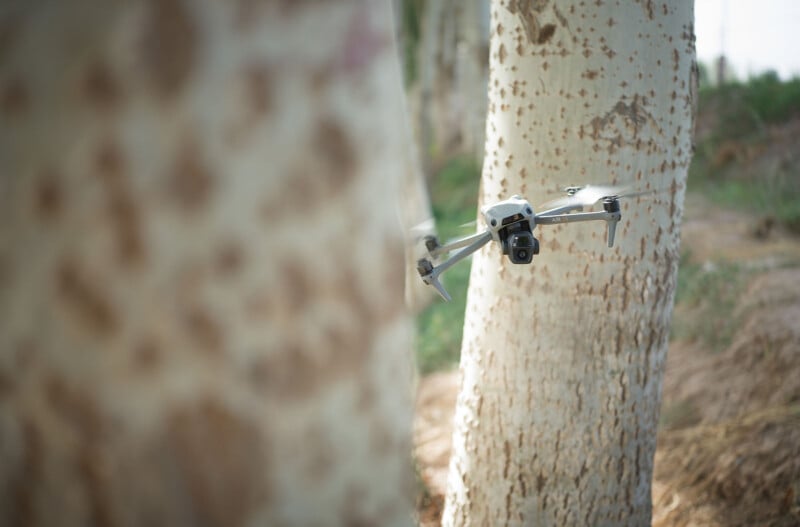 A drone flies close to a tree trunk in a wooded area, partially obscured by the tree. The background shows blurred trees and greenery, suggesting a forest environment.