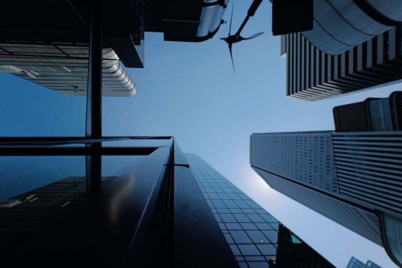 A dramatic upward view of multiple tall skyscrapers against a clear blue sky. The buildings are varied in design, including one with reflective glass and others with distinct architectural features, converging towards a central point.