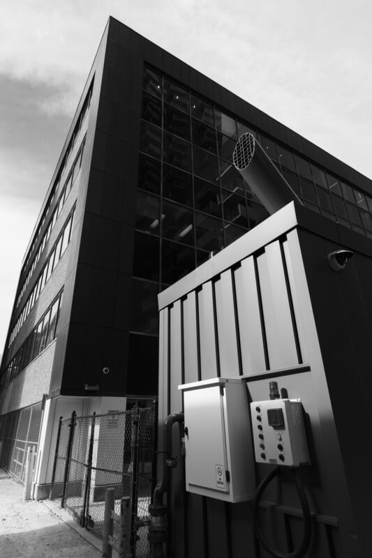 A low-angle black and white photo of a modern building with large glass windows. In the foreground, there is an industrial unit with metal siding, control panels, and a mesh fence. The sky is overcast, adding contrast to the scene.