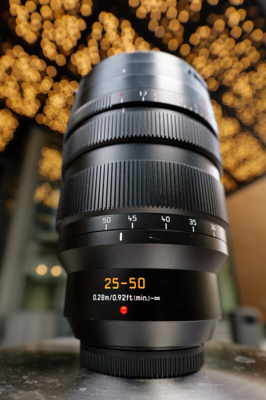 Close-up of a large camera lens with markings showing a focal range of 25-50mm and a minimum focus distance of 0.28m/0.92ft. The background features a bokeh effect with warm, blurred lights.