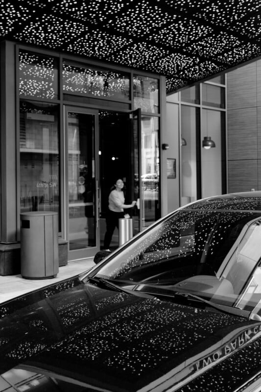 A woman opens a glass door under a canopy filled with small, star-like lights. In the foreground, a black car reflects the lights, creating a shimmering effect. The scene is urban, with modern architecture visible. Black and white photo.