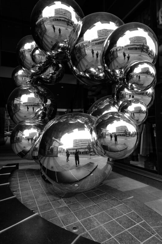 A black and white photo of a reflective sculpture made of interconnected polished spheres, capturing the distorted reflections of nearby buildings and pedestrians. The artwork is displayed outdoors on a tiled surface.