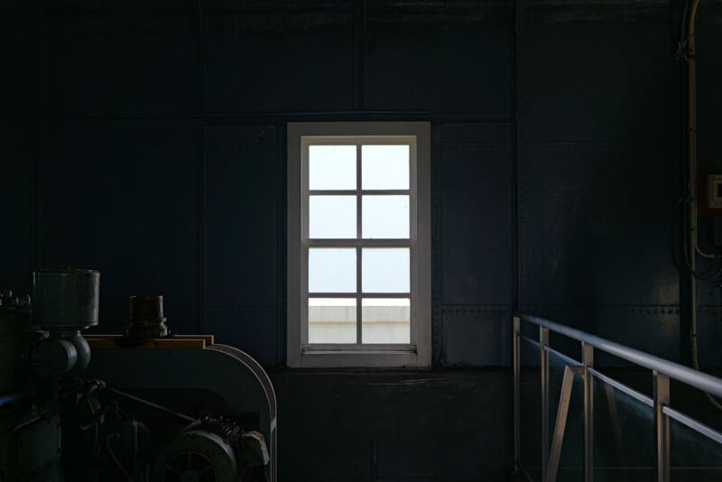 A single window with a grid pattern is centered on a dark industrial wall. The view outside is overexposed, contrasting with the dimly lit room. Metal machinery is partially visible in the foreground on the left.