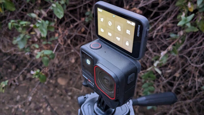 Close-up of an action camera mounted on a tripod outdoors. The camera's screen is tilted upwards, displaying various settings icons. The background consists of out-of-focus foliage.