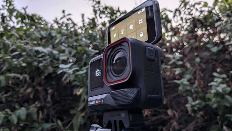 Close-up of an action camera with a touchscreen display tilted upward, showing settings options. The camera is mounted on a tripod against a backdrop of greenery and a cloudy sky.