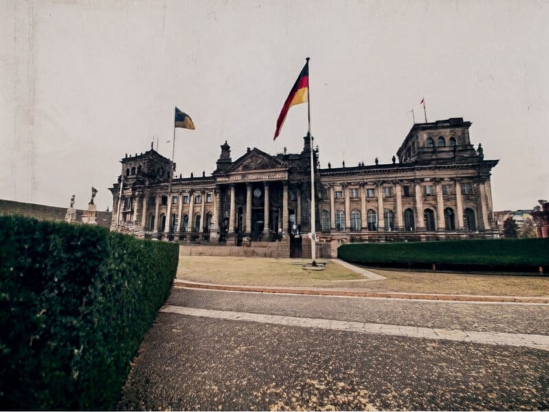 A wide-angle view of the Reichstag building in Berlin, Germany. The German flag is prominently displayed in the foreground. The scene includes well-kept paths and hedges leading up to the historic structure under an overcast sky.