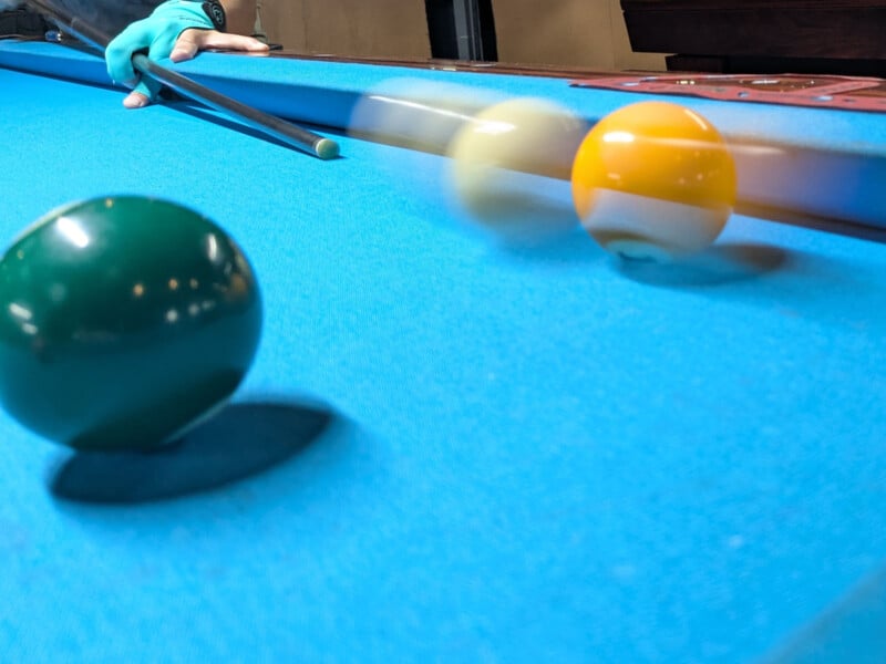 Close-up of a billiard table in action. A person wearing a glove is taking a shot, and the cue ball is in motion, hitting an orange-striped ball while a solid green ball remains stationary.