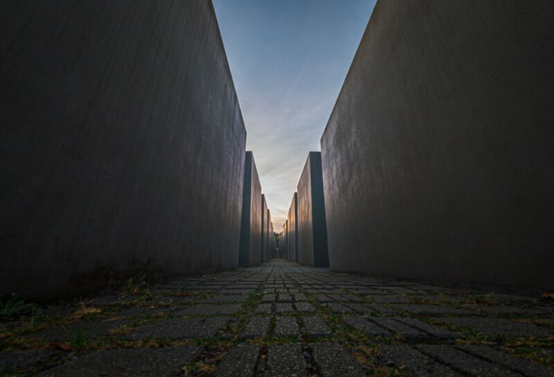 A low-angle view of a pathway flanked by tall, concrete slabs under a clear sky at dusk. The path is paved with stones and the narrow corridor leads towards the horizon, creating a sense of depth and perspective.