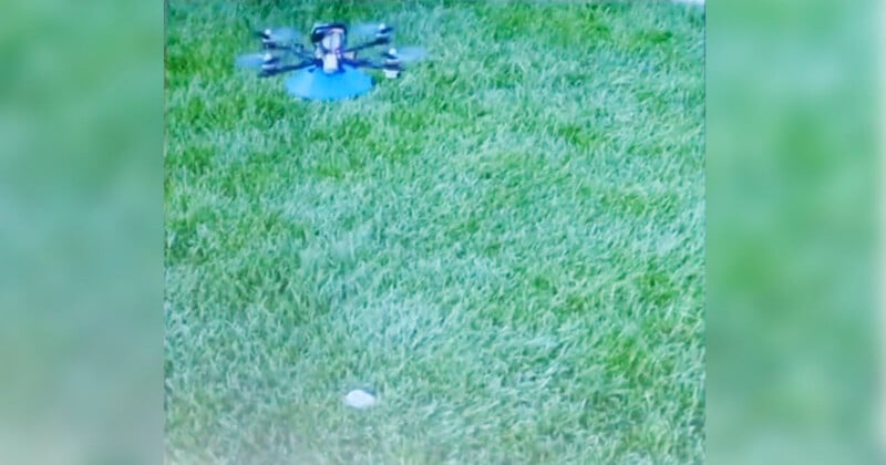 A drone hovers slightly above a well-manicured grassy lawn. Its propellers are visible, showing it in mid-flight. The grass is bright green and evenly cut, suggesting a maintained outdoor setting. The scene is brightly lit, likely indicating daytime.