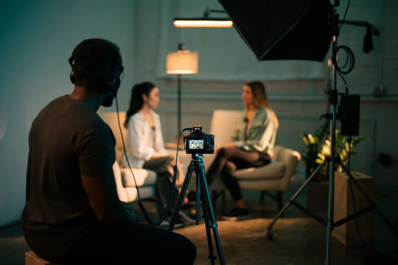 A behind-the-scenes view of a video recording setup. Two women sit in chairs facing each other, engaged in conversation. A camera on a tripod films them, while a person with headphones monitors the scene. Studio lights illuminate the area.