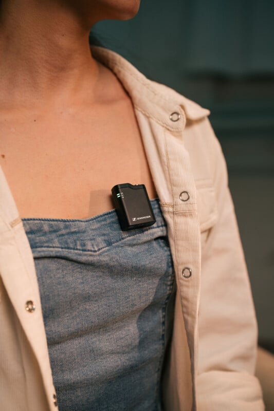 A person wearing a denim top and a light-colored open jacket has a small black wireless microphone clipped to their chest. The background is softly blurred.