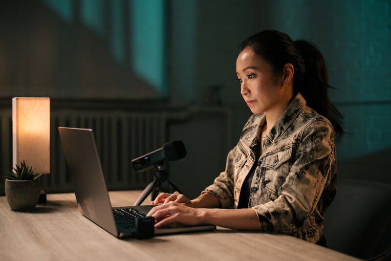 A woman sits at a desk with a laptop, typing while looking at the screen. A microphone is set up on the desk beside her. A softly lit lamp and a small potted plant are also on the table. The room has a dim, moody ambiance.