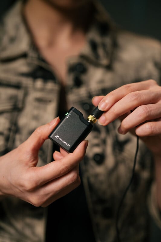 A person wearing a denim jacket holds a small black electronic device with a cable. They are adjusting the plug on the device, which has a logo and indicator lights. The focus is on the hands and the device.