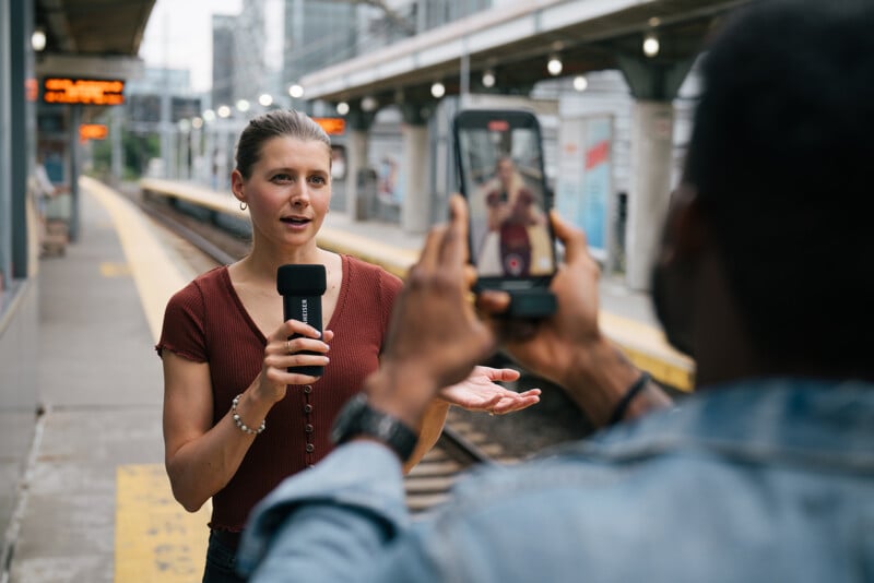 A woman holding a microphone is talking while a person films her with a mobile phone at a train station platform. Blurred trains and digital signs are visible in the background under a covered walkway.