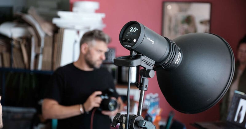 A close-up of a studio light in focus, with a digital display reading 3.0. In the blurred background, a person holds a camera, suggesting a photography shoot in progress. The setting includes various studio equipment and a red wall.