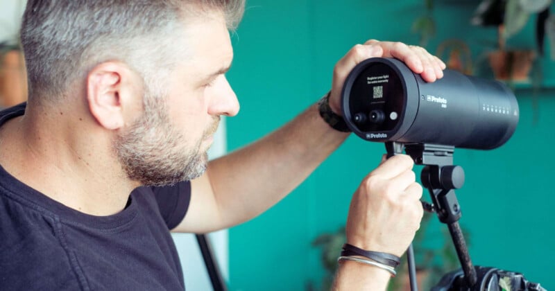 A man adjusts a professional camera light on a stand. He is wearing a black shirt and has short hair and a beard. The background is teal with some plants visible.