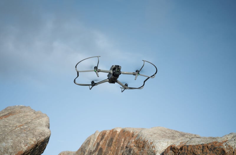 A drone with protective guards hovers in a clear blue sky above large rocks. The drone's camera is visible, and the scene is illuminated by natural daylight.