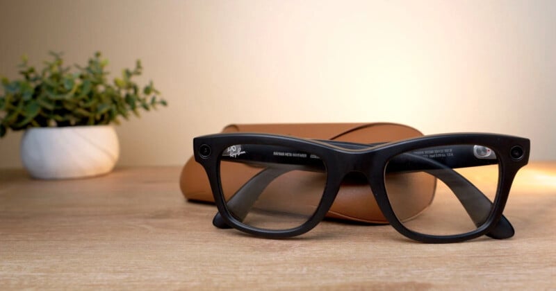 A pair of black-framed smart glasses is displayed on a wooden surface in front of a brown glasses case. In the background, there is a small potted plant with green leaves situated on the left side.