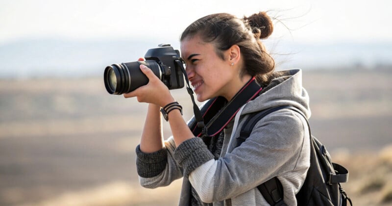 A young woman with a backpack is smiling and taking a photograph with a DSLR camera outdoors. She's wearing a gray hoodie and has her hair tied up in a bun. The background is a blurred landscape.