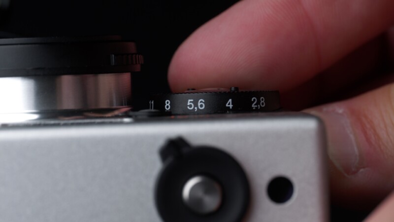 Close-up of a person's hand adjusting the aperture dial on a camera lens. The dial shows aperture settings: 11, 8, 5.6, 4, and 2.8. The camera body is silver with a black button visible in the foreground.