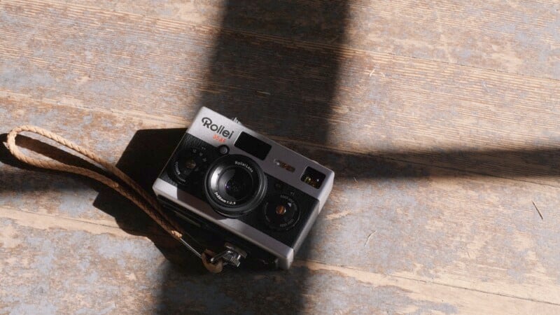 A vintage Rollei 35 film camera with a brown strap is placed on a wooden floor, partially illuminated by sunlight streaming through a window, casting an intersecting shadow.