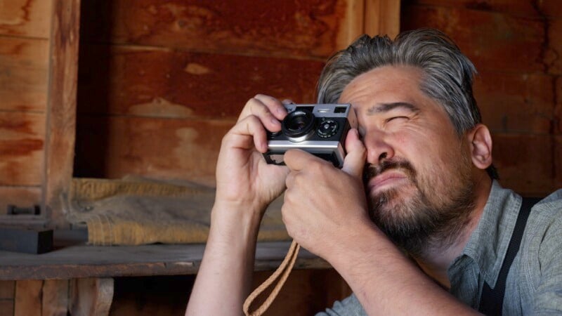 A person with gray hair and a beard is inside a wooden cabin, holding a vintage camera to their eye and appearing focused on taking a photo. They wear a light gray shirt, and some fabric is on the wooden surface in the background.