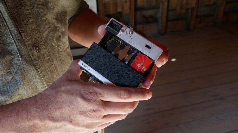 A person loads a film cartridge into a vintage Polaroid camera. They are wearing a green button-up shirt. The background is dimly lit with wooden walls and flooring, suggesting an indoor setting.