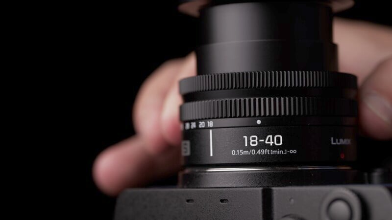 Close-up of a person's hand adjusting the zoom ring on a Lumix camera lens showing focal length markings from 18 to 40mm against a black background.