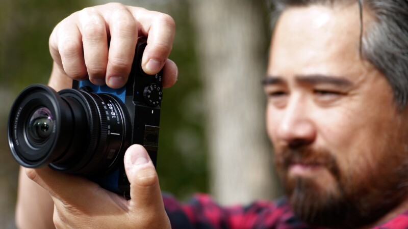 A person with a beard, wearing a red plaid shirt, is holding a camera up to their eye, seemingly about to take a photo. The background is blurred, suggesting an outdoor setting.