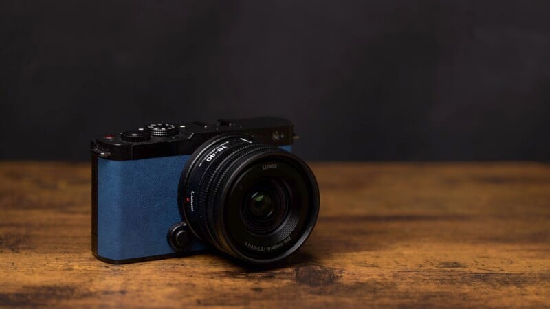 A blue and black digital camera with a lens cap is placed on a wooden surface against a dark background.