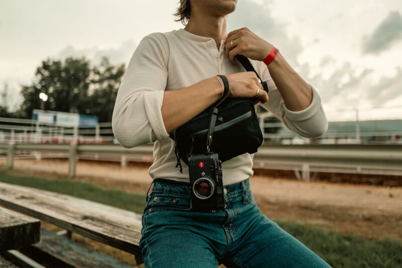 A person in a white long-sleeve shirt and jeans sits on a bench, holding a fanny pack. They have a camera attached to the pack and wear a red bracelet. A racetrack and cloudy sky are in the background.